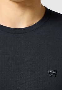 Primer plano de una persona que lleva una camiseta negra de cuello redondo lisa con un pequeño parche negro del logo de Wrangler en el pecho.