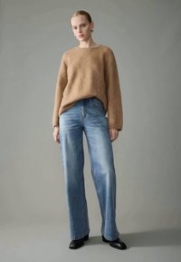 Suéter de malha fluffy castanho claro emparelhado com calças de ganga largas azuis desbotadas. Modelo veste botas pretas de salto, posando contra um fundo neutro.