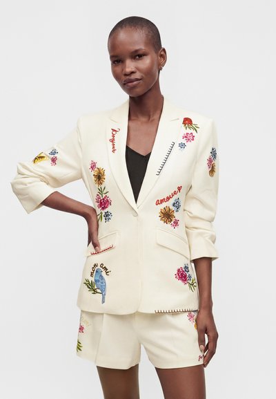 Personne portant un blazer crème et un short avec broderies colorées de fleurs, d'oiseaux et de mots français, posant la main sur la hanche devant un fond clair.