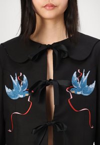 Giacca nera con un ampio colletto, caratterizzata da due ricami di uccelli blu e dettagli rosa, chiusa con nastri neri.