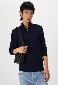 Suéter de punto navy con cuello alto y cierre de cremallera. Combinado con unos vaqueros azul claro y un pequeño bolso cruzado negro.