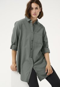 Rohelise värvusega nööbitav särk rinnatasku, rullitavate varrukatega ja lõdva äärega, valmistatud pehmest tekstuuriga materjalist. Allpool on seljas mustad püksid.