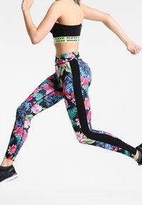 Leggings à motifs floraux aux couleurs vives avec des panneaux latéraux noirs, en matériau extensible. Associés avec un soutien-gorge de sport noir.