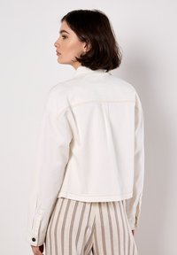 Weiße Jeansjacke mit einer verkürzten Passform, sichtbaren Nähten und Knopfakzenten an den Ärmeln, kombiniert mit gestreiften beige- und weißen Hosen.