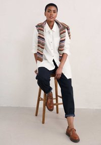 Biała koszula z długim rękawem z podwiniętymi rękawami, niebieskie dżinsy, wielokolorowa kamizelka zarzucona na ramiona oraz brązowe buty.