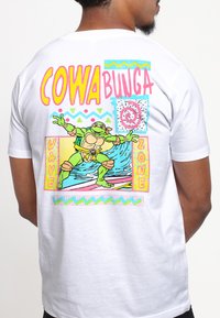 Hvid t-shirt med et farverigt tegneserie-skjoldpaddedesign. Tekst "COWABUNGA" og "WAVE ZONE" i lys pink, gul og blå.