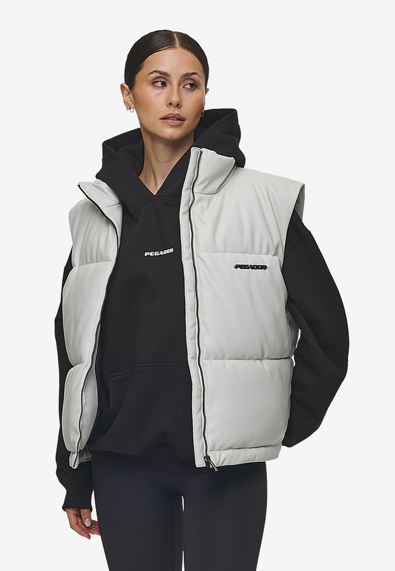Lys grå quiltet puffer vest med høy krage, båret over en svart hettegenser. Har frontlommer og subtil logo-detaljering.