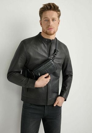 Mężczyzna w czarnej skórzanej kurtce i ciemnych dżinsach trzyma czarną torebkę przez ramię na jednolitym jasnym tle.