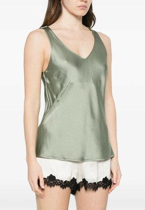 Donna che indossa un top senza maniche in raso verde chiaro e pantaloncini in seta bianca con rifiniture in pizzo nero, in piedi contro uno sfondo bianco semplice.