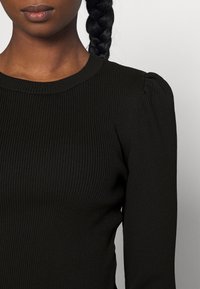Haut en tricot côtelé noir avec un col rond et des manches légèrement bouffantes, présentant une coupe ajustée et une texture lisse.