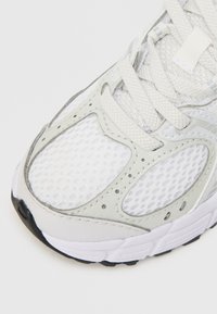 Zapatilla deportiva gris claro con parte superior de malla, acentos texturizados y cordones blancos. La suela exterior es negra con un dibujo multidireccional.