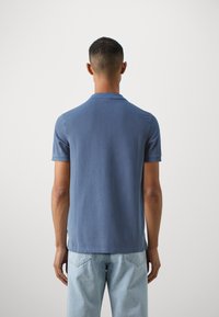 Modra kratka polo majica iz mehkega bombaža. Ima klasičen ovratnik in ravno spodnje rob. Zadnji pogled, v kombinaciji s svetlo modrimi jeans hlačami.