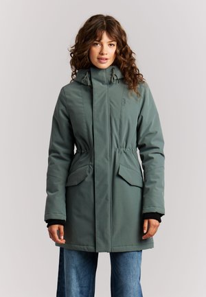 Vrouw met krullend haar, gekleed in een groene winterjas met capuchon en klepzakken en blauwe spijkerbroek, staand tegen een effen achtergrond.