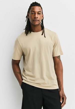 Homme avec des dreadlocks aux épaules, portant un t-shirt beige et un pantalon noir, debout avec une main dans la poche sur fond blanc.