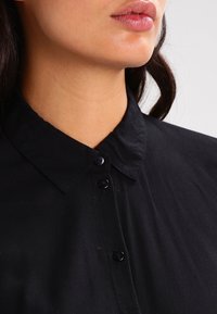 Schwarz geknöpfte Bluse aus glattem Stoff, mit einer Knopfleiste und dunklen Knöpfen. Der Kragen und die Textur sind deutlich sichtbar.