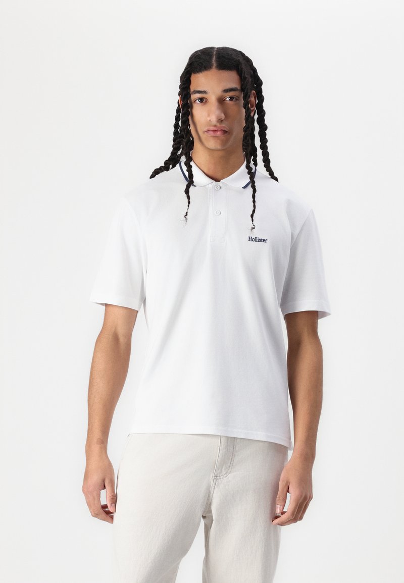 Bílá polokošile vyrobená z bavlny, s tříknoflíkovou légou a modrým vyšíváním. Logo je na levé hrudi. Model má na sobě světlé kalhoty.