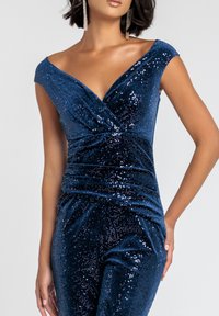 Jumpsuit blu con paillettes, scollatura a V profonda, maniche a cappuccio e dettagli arricciati in vita. Il tessuto ha un aspetto morbido e testurizzato.