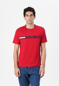 Rode katoenen T-shirt met korte mouwen, voorzien van een horizontale zwarte, witte en marineblauwe streep over de borst met de tekst 'TOMMY HILFIGER'.