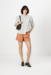 Floral print shirt in light blue and white, with puffed sleeves; paired with orange shorts; accessorized with a small brown handbag and dark shoes.
