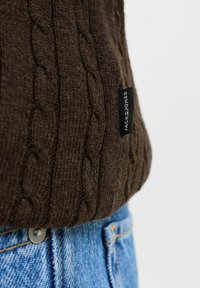 Pulover maro tricotat cu model texturat, având un etichetă neagră inscripționată "JACK & JONES," asortat cu blugi din denim albastri.