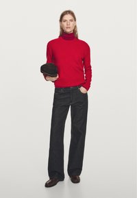 Červený rolák spojený s tmavě modrými širokými džíny. Drží tmavě hnědou čepici s jednoduchým designem a texturovanou látkou.