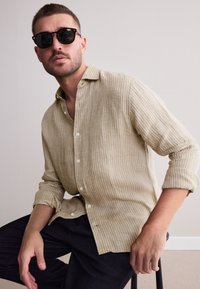 Camicia di lino a righe color senape con chiusura a bottoni e maniche arrotolate, abbinata a pantaloni neri. Modella con occhiali da sole rotondi neri.