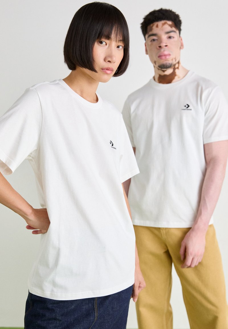 Camisetas de algodón blancas con mangas cortas, que presentan un pequeño logo negro en el pecho. El modelo lleva puestos jeans azules; el segundo modelo lleva pantalones amarillos.