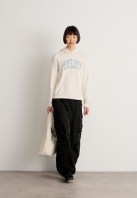 Person wearing a cream hoodie with "GANT 1949 NEW HAVEN" text, black cargo pants, holding a white tote bag, standing against a plain white wall.