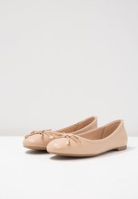 Coppia di ballerine beige in pelle con punte arrotondate, piccoli fiocchi e dettagli in pizzo con punte dorate su una superficie bianca.