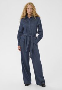 Ženska z dolgimi kodrastimi blond lasmi nosi temno modro jeans kombinezon z zavezaným pasom in črnimi čevlji na enobarvni beli podlagi.