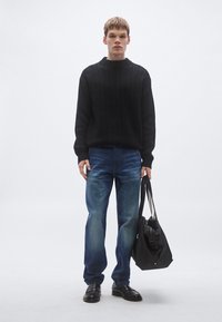 Musta kootud kampsun, sinised teksapüksid, mustad nahast kingad ja must kott rihmaga. Modell seisab ühtlase valge tausta ees.