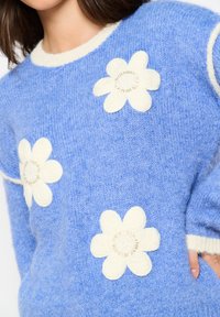 Sininen neulepaita, jossa on valkoiset kukkakoristeet ja teksturoitu pinta. Ominaisuuksiin kuuluu pyöreä pääntie ja kontrastiväriset hihansuut.