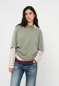 Tricou verde din bumbac supradimensionat, purtat peste o cămașă în dungi cu guler; asortat cu blugi albaștri uzați pentru un look casual.