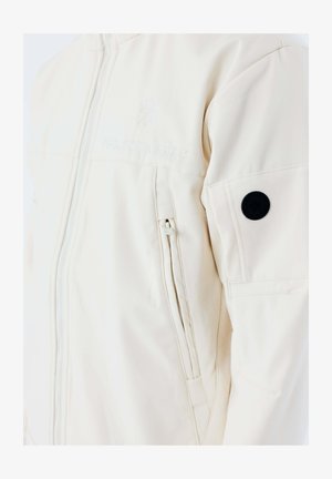 Off-white jas met ritssluiting aan de voorkant, ritssluiting aan de zijkant, geborduurd logo op de borst en een zwarte ronde patch op de bovenarm.