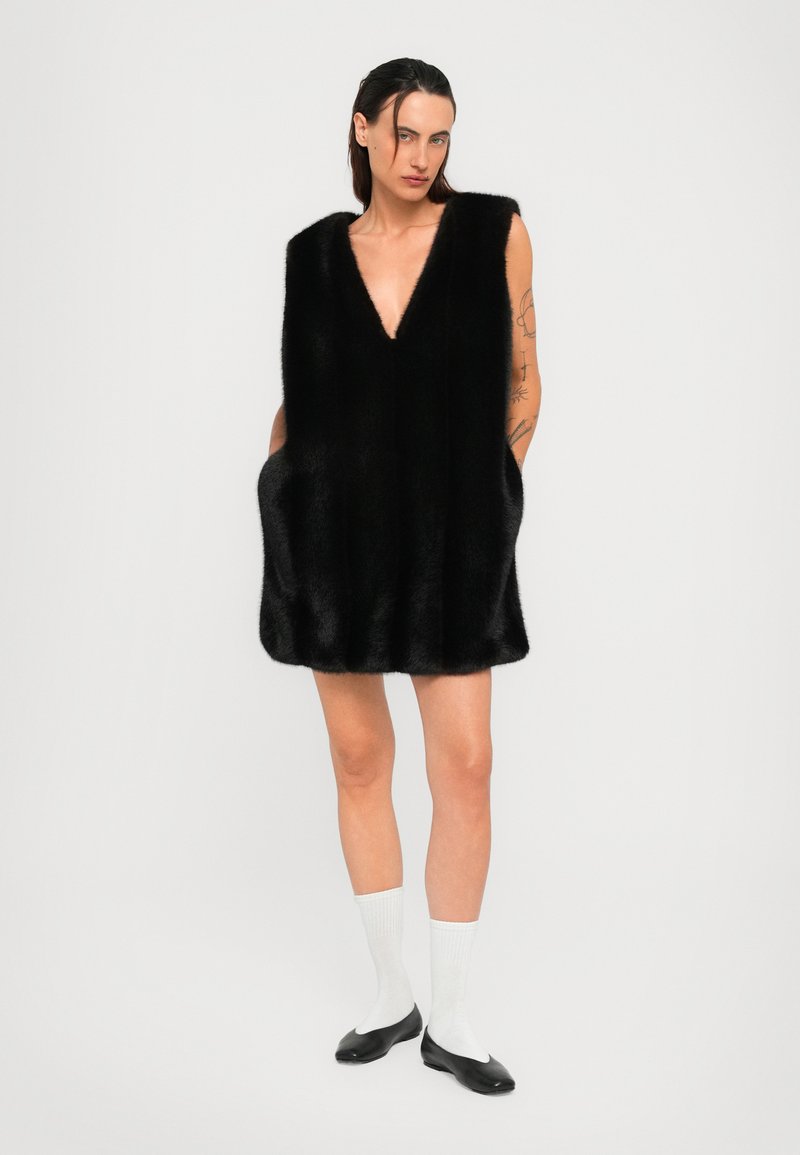 Person wearing a sleeveless black fur dress with pockets, white mid-calf socks, and black ballet flats standing against a plain background.