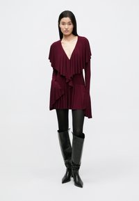 Rochie burgund cu volane suprapuse, decolteu adânc în V și mâneci lungi. Purtați cu cizme negre cu toc stiletto până la genunchi și colanți opaci.