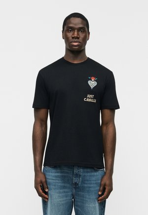 Muž nosiaci čierne tričko Just Cavalli s kvetinovým srdcovým dizajnom a textom na hrudníku, v kombinácii s modrými džínsami, stojaci pred jednoduchým pozadím.