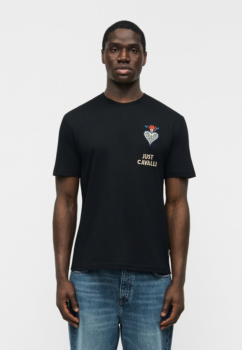 Άνδρας που φοράει μαύρο t-shirt Just Cavalli με σχεδιασμό λουλουδιού σε σχήμα καρδιάς και κείμενο στο στήθος, σε συνδυασμό με μπλε τζιν, στέκεται μπροστά σε μια απλή φόντο.