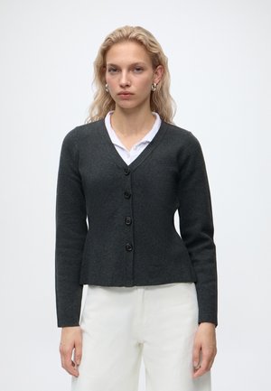 Mujer rubia llevando pendientes de aro plateados, cárdigan gris oscuro abotonado sobre una camisa blanca con cuello y pantalones blancos, de pie frente a un fondo liso.