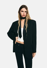 Hosszú szőke hajú nő túlságosan nagy fekete zakót, fekete nadrágot, fehér crop topot és fekete nyakkendőt visel egyszínű háttér előtt.