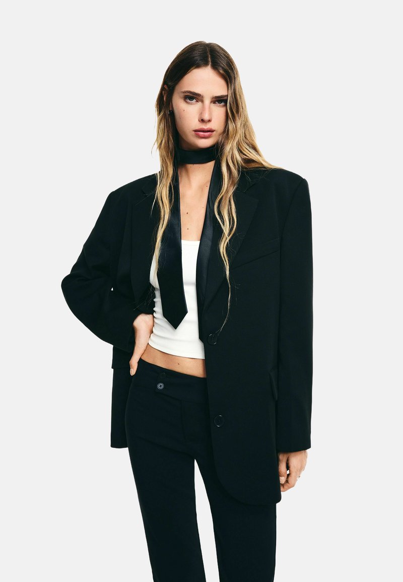 Hosszú szőke hajú nő túlságosan nagy fekete zakót, fekete nadrágot, fehér crop topot és fekete nyakkendőt visel egyszínű háttér előtt.