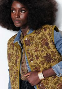 Gilet à motif floral jaune-vert avec des manches en denim, associé à une blouse à motif. Bracelet en métal doré au poignet.