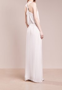 Kobieta ubrana w białą, bezrękawnikową, plisowaną suknię maxi z plecami w kształcie skrętu, stojąca na beżowym tle.