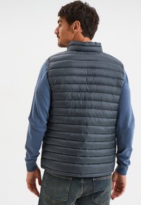 Marineblauer Steppweste mit hohem Kragen, gesteppter Optik und glatter Textur, getragen über einem blauen Langarmshirt und Jeans.