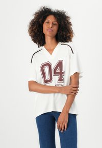Weißes V-Ausschnitt-Sporttrikot mit maroonfarbener "04"-Druck und schwarzen Schulterakzenten, getragen mit blauen Jeans, mit lockeren kurzen Ärmeln.
