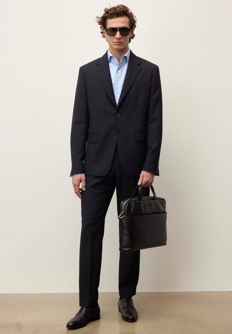 Homme en costume noir, chemise bleu clair, lunettes de soleil noires, tenant une mallette en cuir noire, debout devant un fond clair uni.