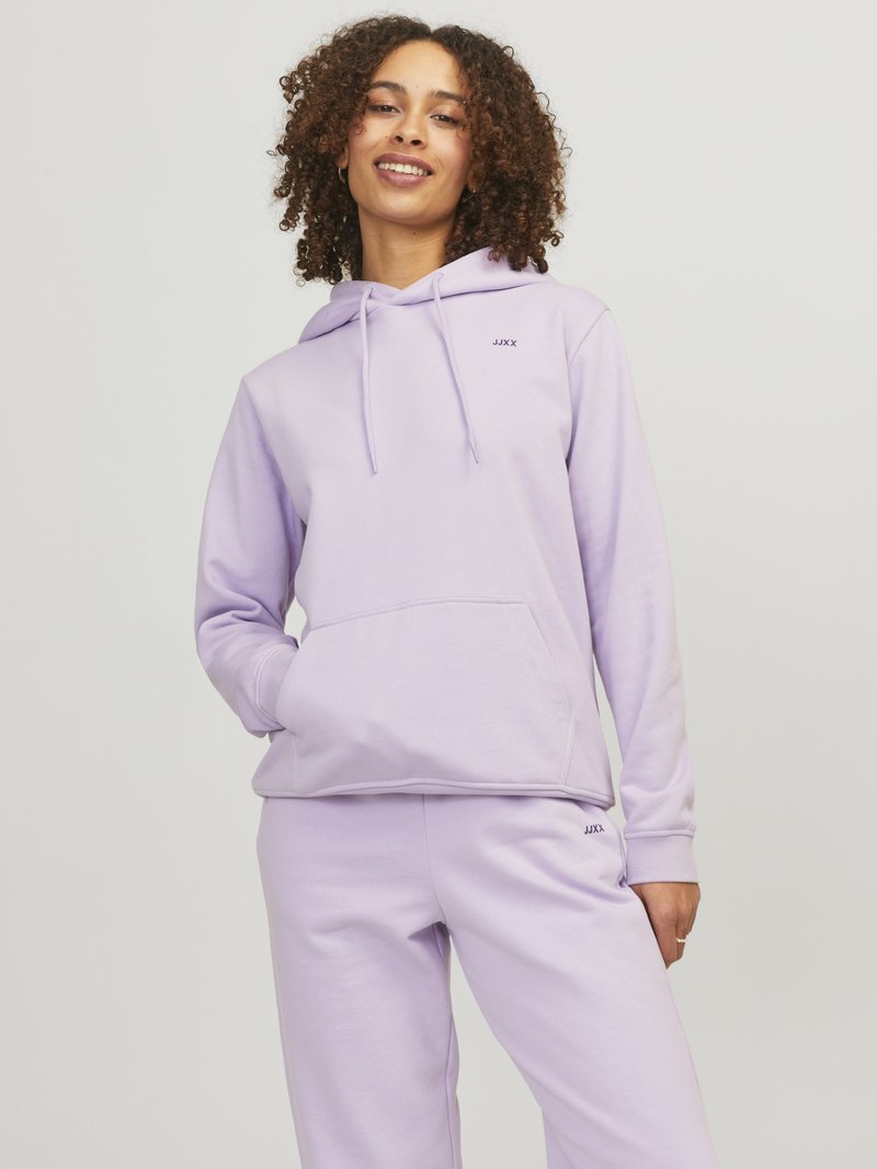Vrouw met krullend haar die een lichtpaarse hoodie en bijpassende joggingbroek draagt, staand met één hand in de zak, glimlachend tegen een effen achtergrond.