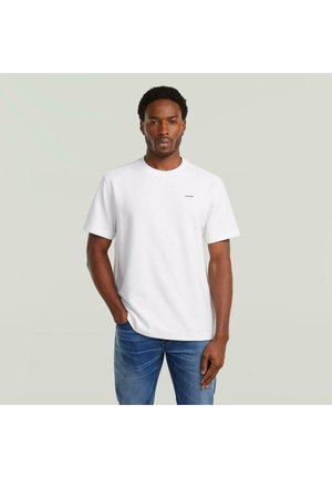 Homme aux cheveux courts et bouclés portant un t-shirt blanc uni à manches courtes et un jean bleu, debout devant un fond gris clair.