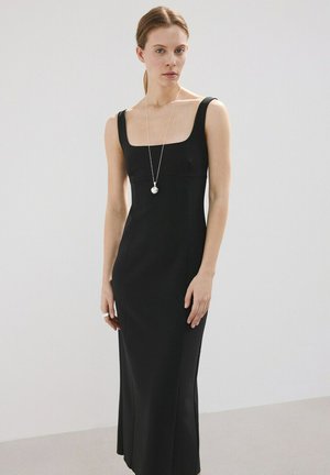 Femme à la peau claire et cheveux attachés portant une longue robe noire sans manches et un collier pendentif, debout devant un fond clair uni.