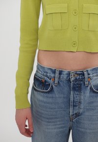 Bright green, cropped cardigan with two front pockets and ribbed cuffs paired with faded blue jeans featuring orange stitching and a classic button closure.
